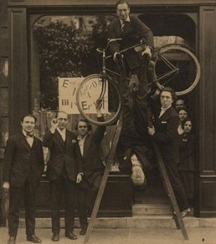 Vernissage de l'exposition Ernst à la galerie Au Sans Pareil, Paris (1921). De gauche à droite : René Hilsum, Benjamin Péret, Serge Charchoune, Philippe Soupault en haut de l'échelle, Jacques Rigaut (à l'envers) et André Breton.