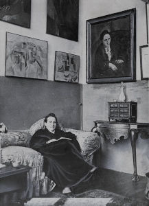 Sur cette photographie de 1930, Gertrude Stein est assise dans son studio parisien. Derrière elle se trouvent quelques tableaux de sa collection d'art moderne, dont le portrait que Pablo Picasso a peint d'elle en 1906.