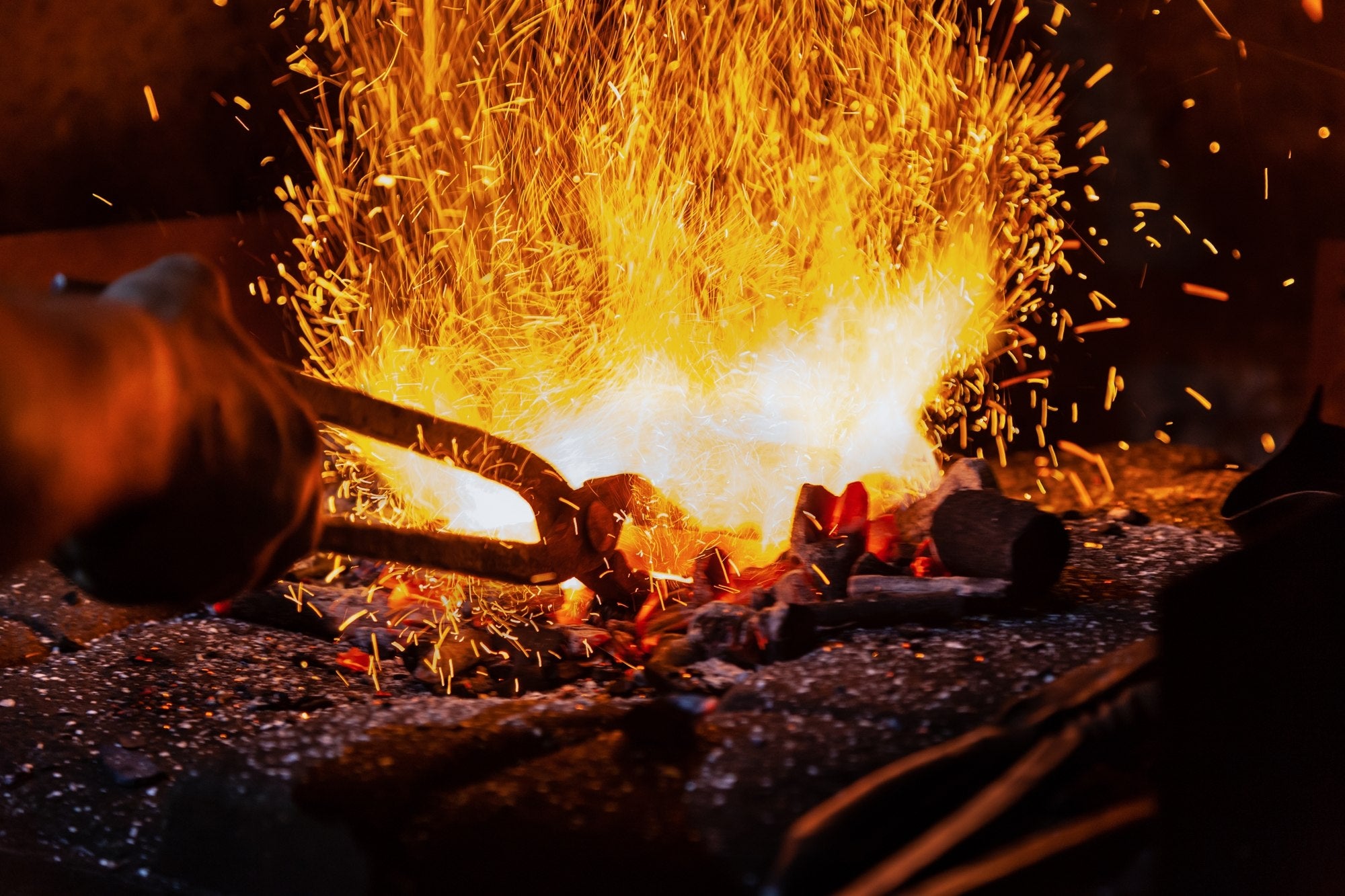 The hands of the blacksmith, unrecognizable, are shown preparing the metal for forging, with sparks flying in the background