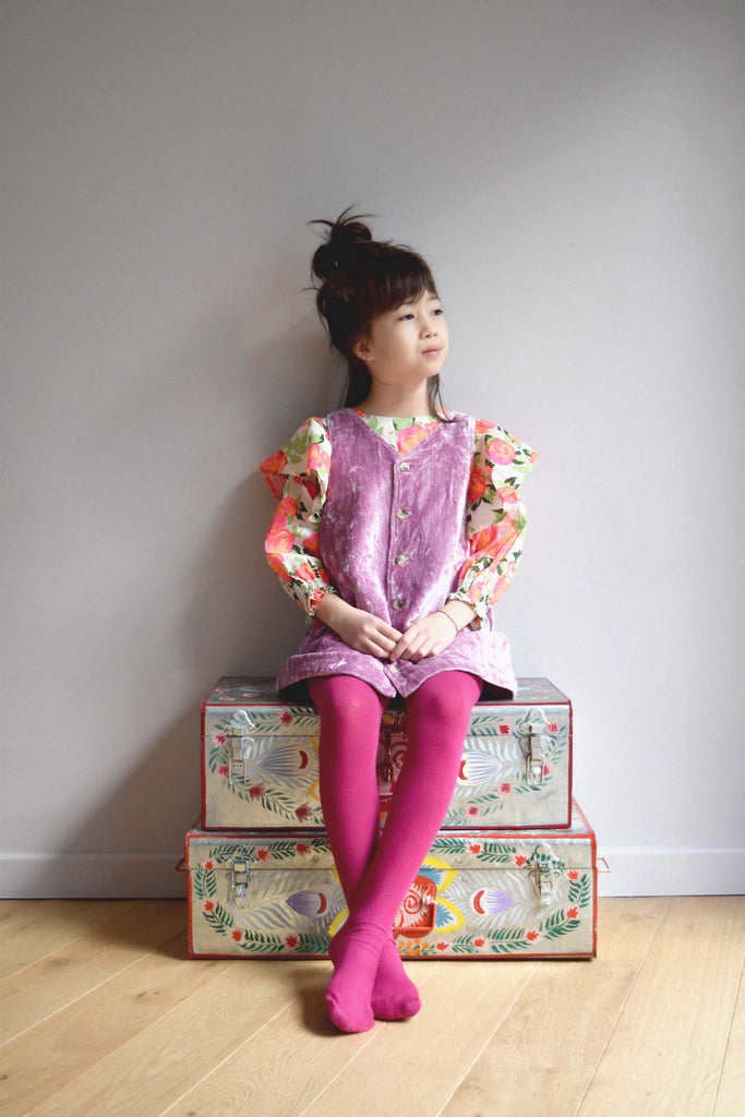 lulaland Fall No.15 Mirage collection. Girl wearing a flowery blouse and a lilac jumper sitting on adorned trunk