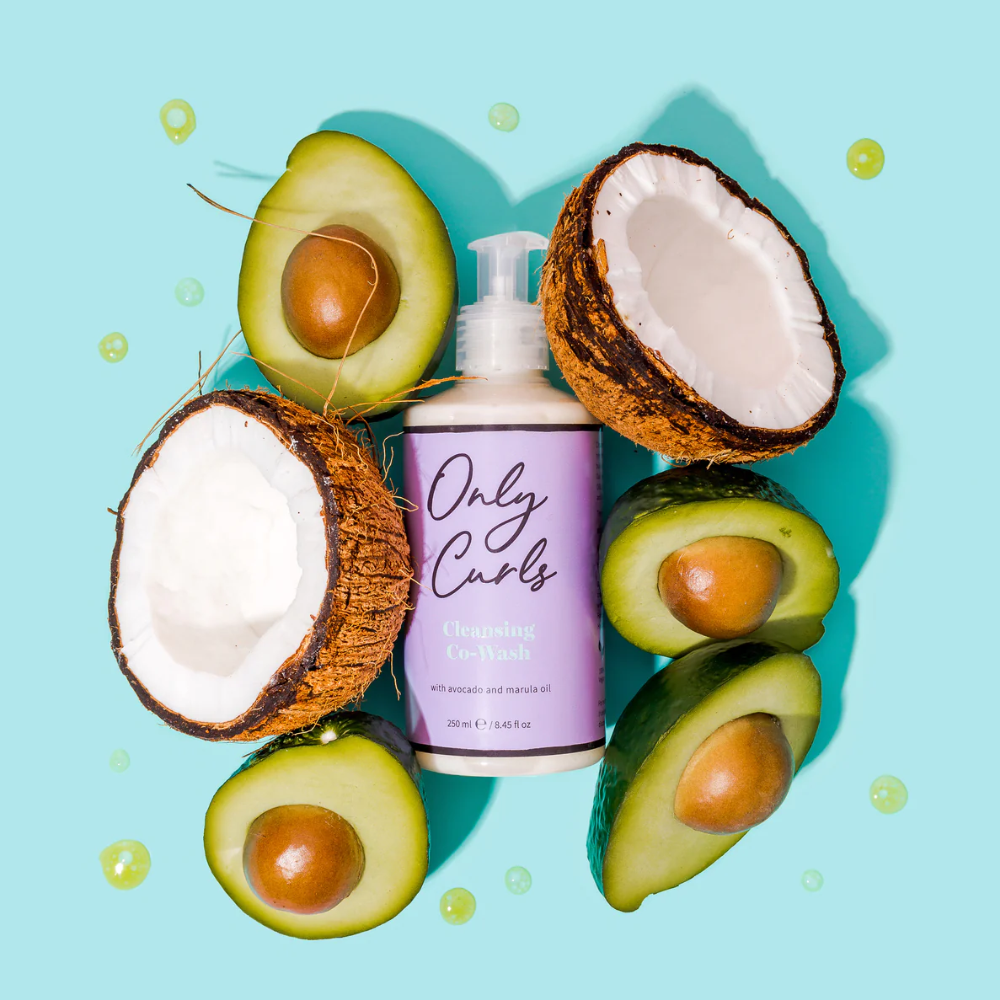 Only Curls Cleansing Co-Wash fles omringd door verse avocado's en kokosnoten tegen een turquoise achtergrond, benadrukkend de natuurlijke ingrediënten.