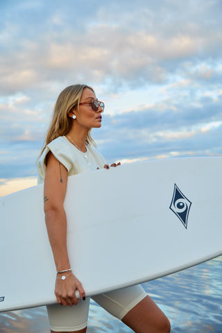 Woman in white clothes and silver bracelets and silver earrings holding a surfboard