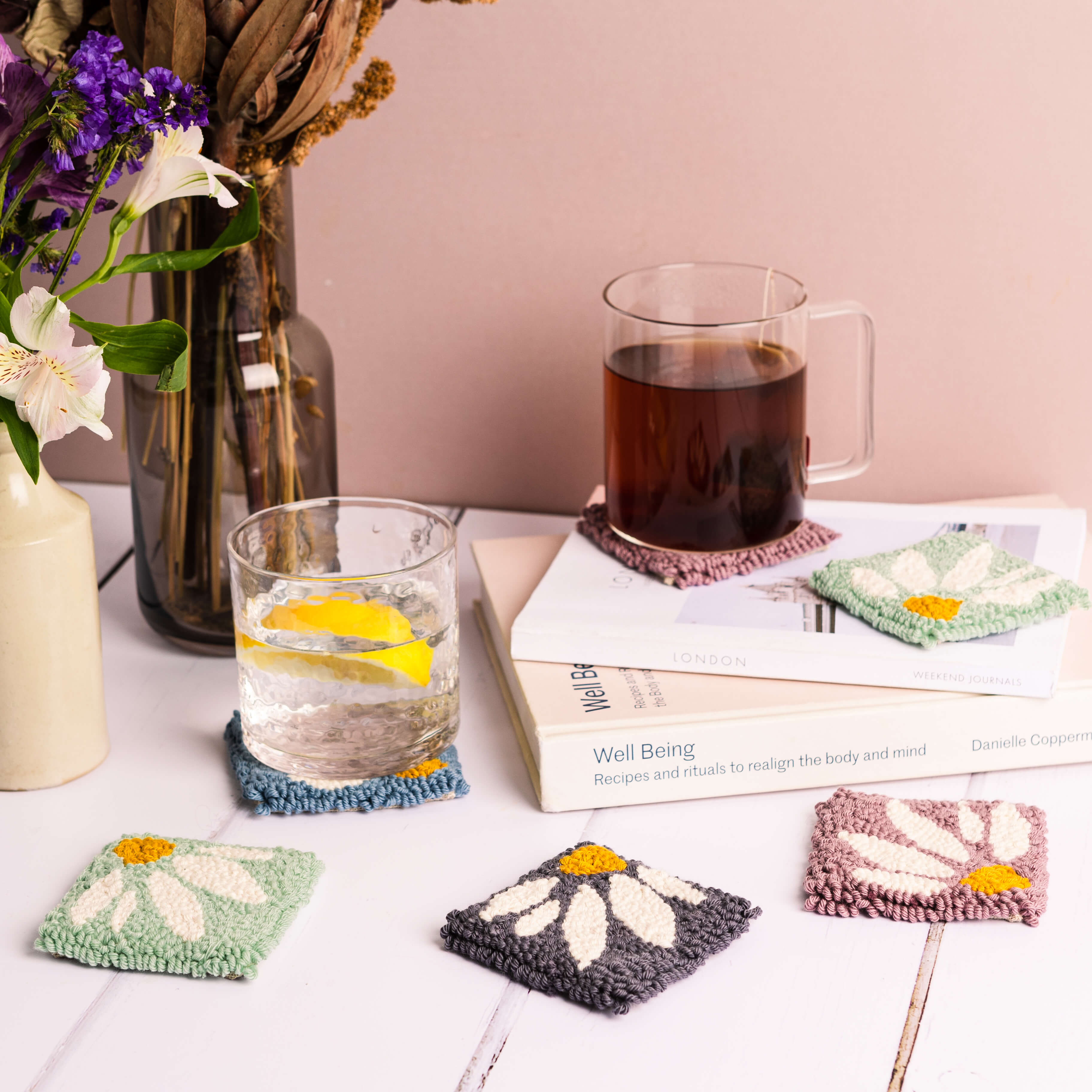 Daisy punch needle coasters with a pink background, stack of books with tea and water resting on a coupl eof coasters, flowers just coming into shot
