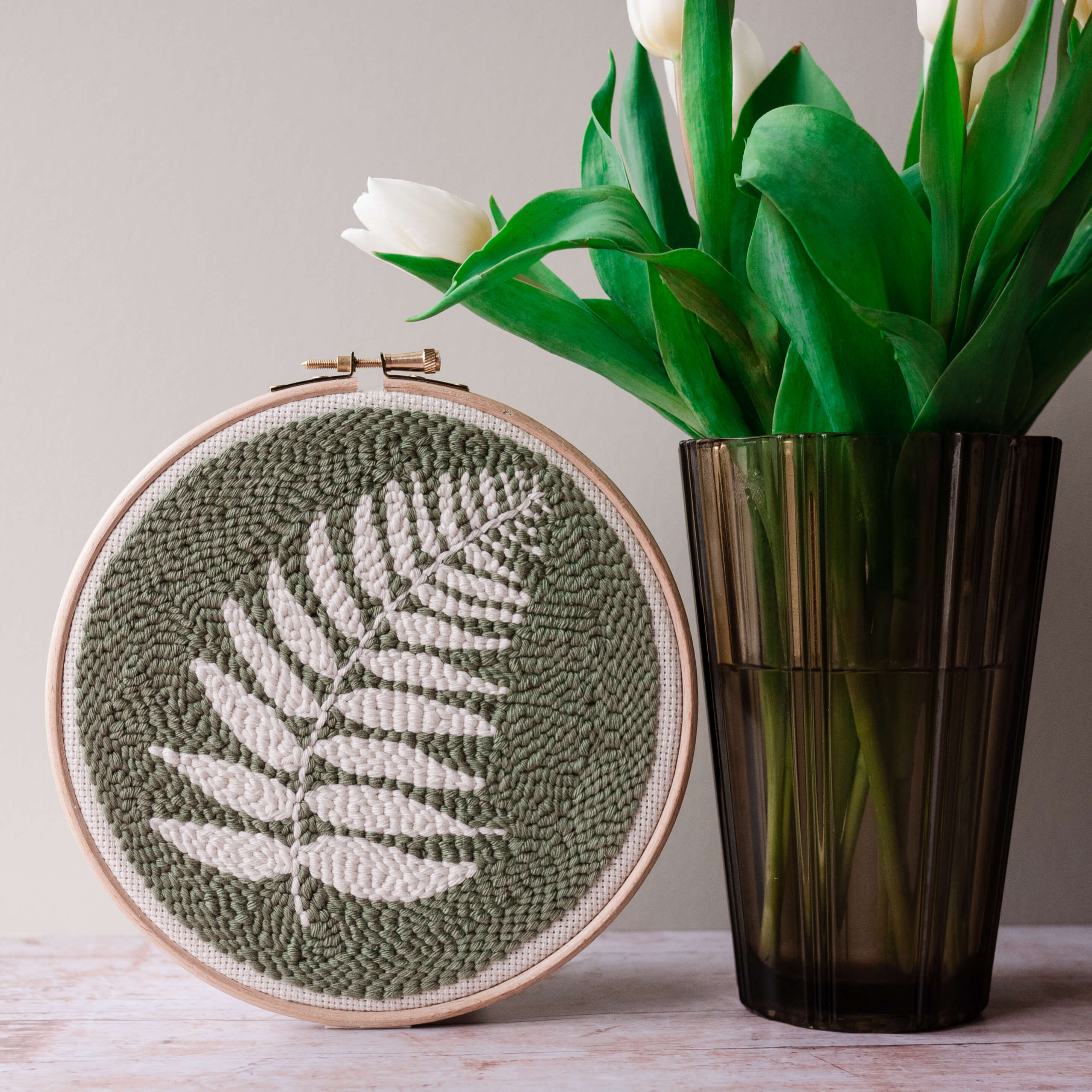 Fern finished punch needle hoop propped up next to a brown glass vase with white tulips in