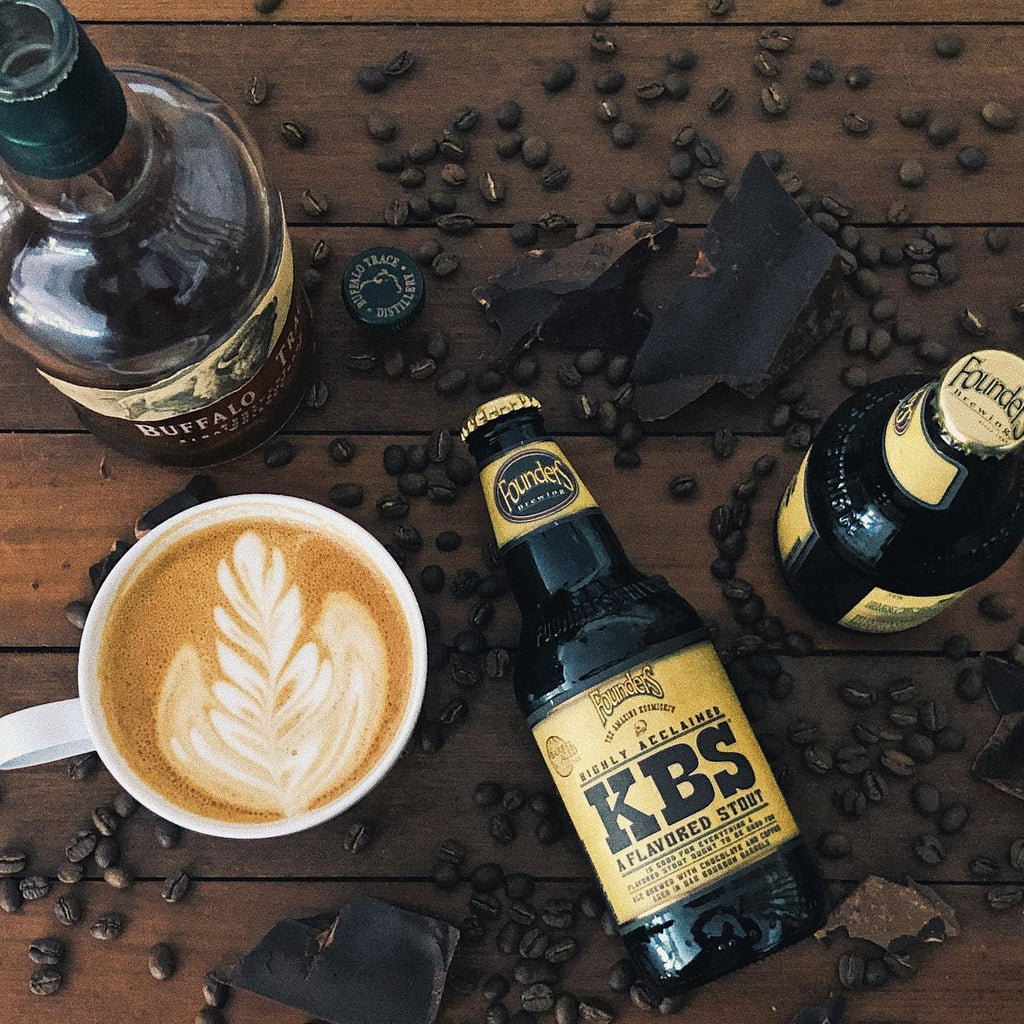 Photo of a cappuccino and beer bottles on a table with chocolate and coffee beans scattered around it. 