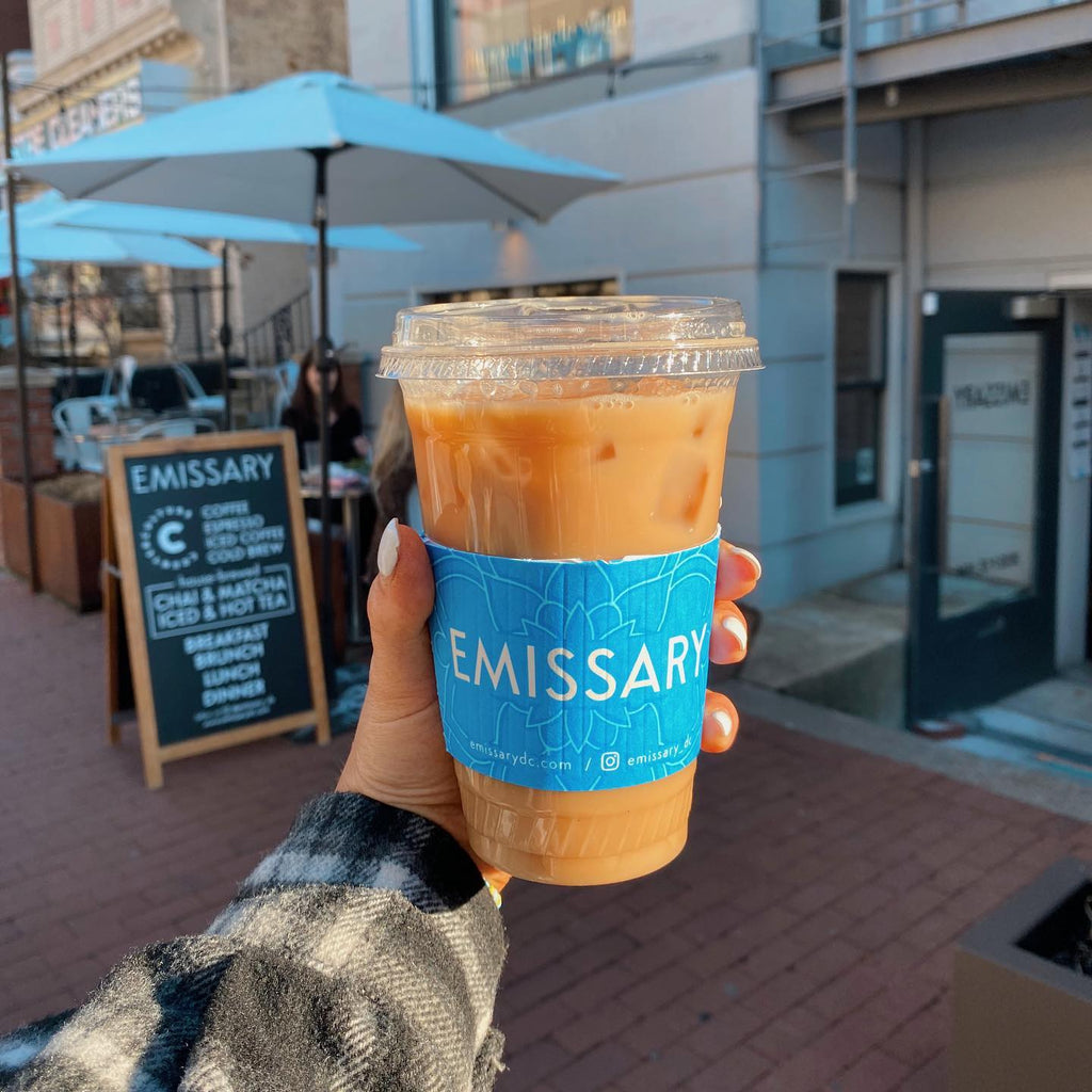 Photo of a hand holding a to-go coffee cup with a sleeve that says, "Emissary."