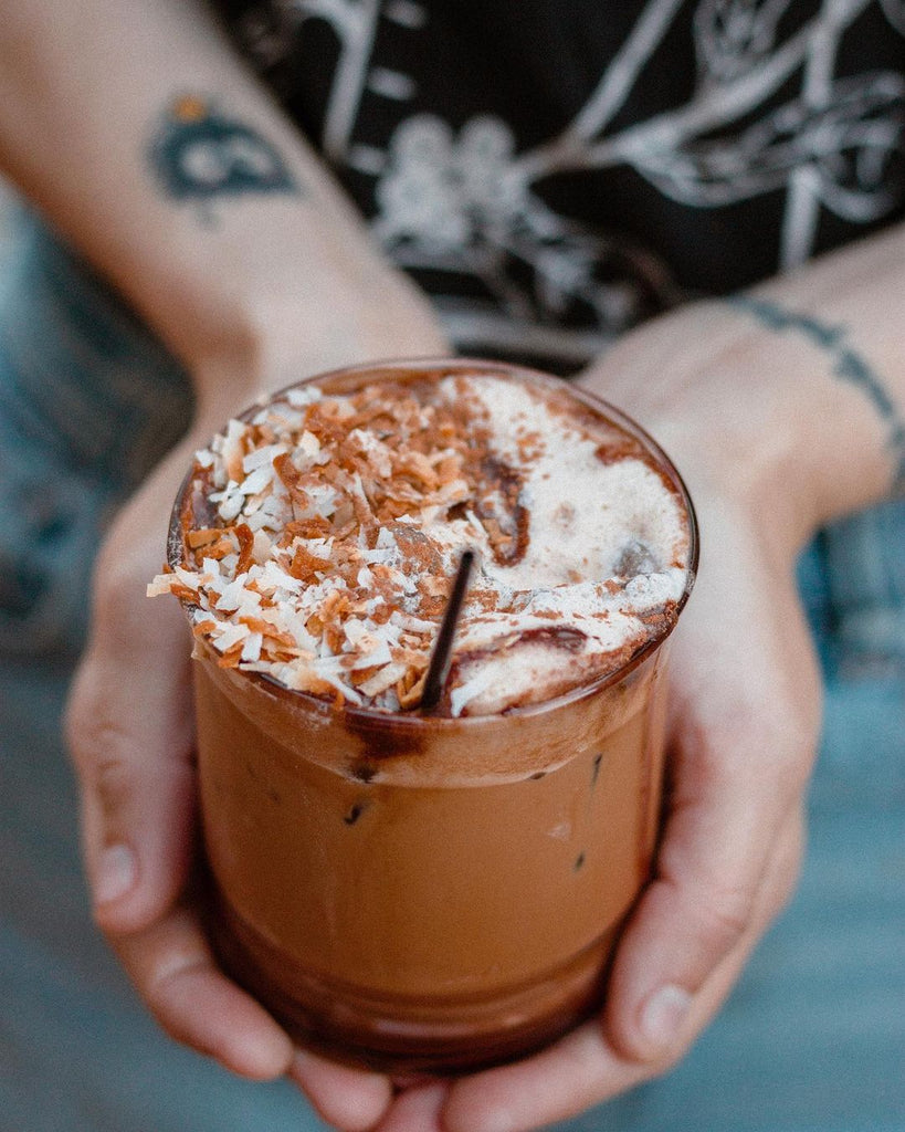 Photo of a glass cupped in the palms of hands. The cup is filled with coffee, ice, and topped with toasted coconut. 