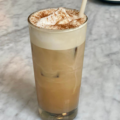 Photo of a glass filled with coffee and ice, a large section of foam at the top, and a straw in it. The drink is sitting on a marble counter. 