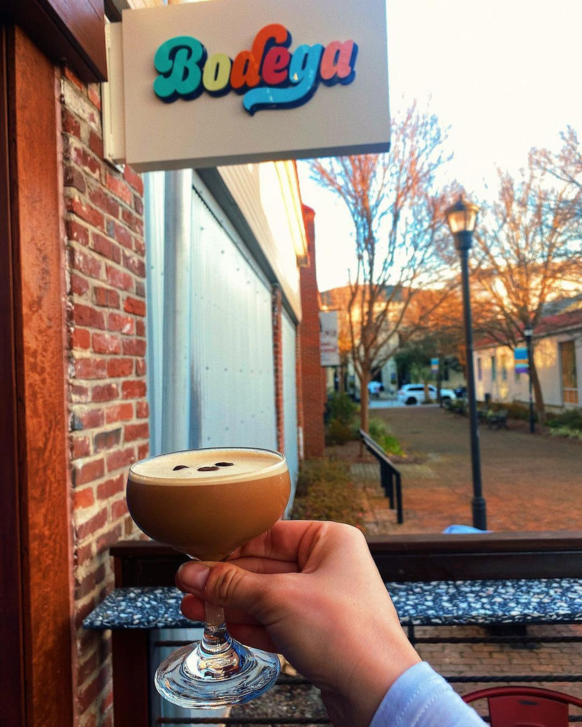 Photo of a hand holding out an espresso martini in front of a sign that says "Bodega"