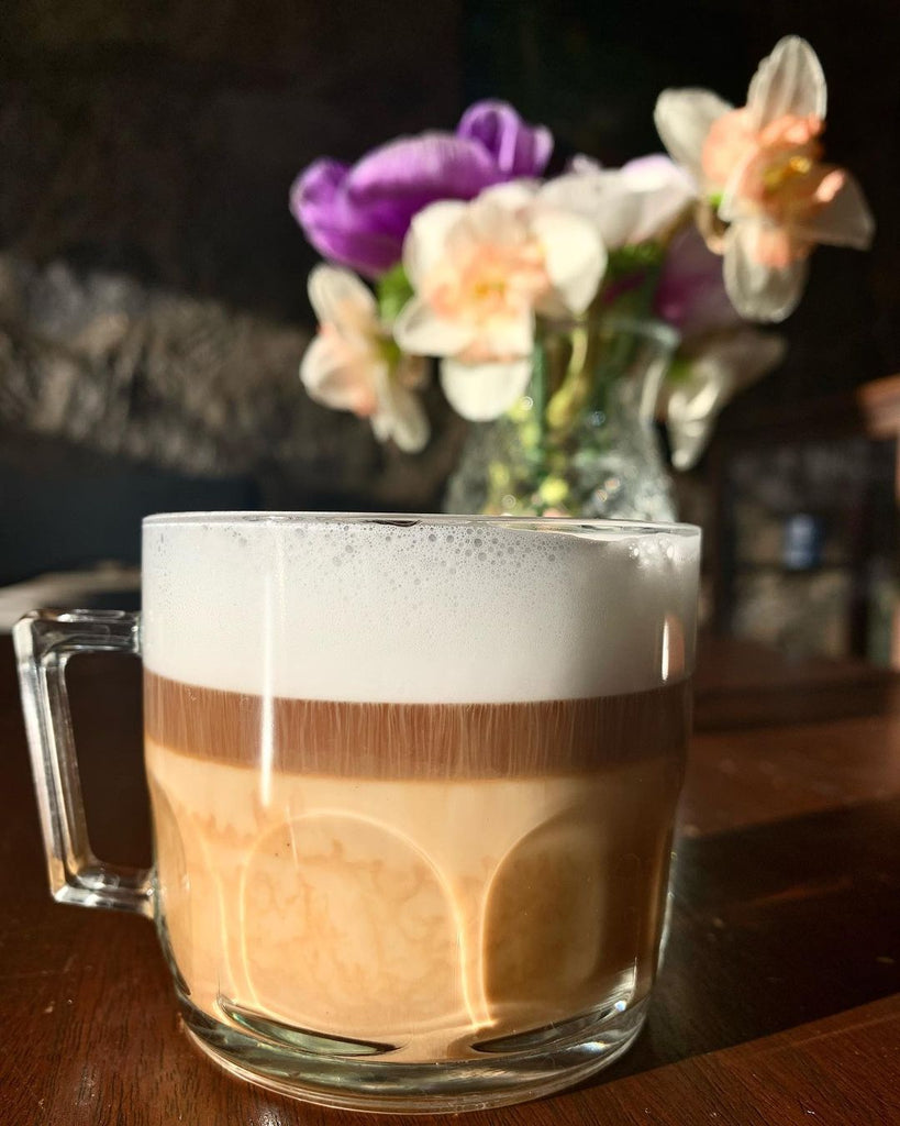 Photo of a layered coffee drink in a glass in front of a vase of spring flowers.