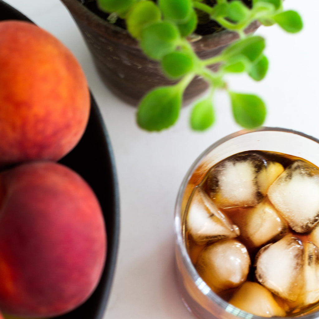 Close up of a glass of iced cold brew next to a bowl of peaches and a plant.