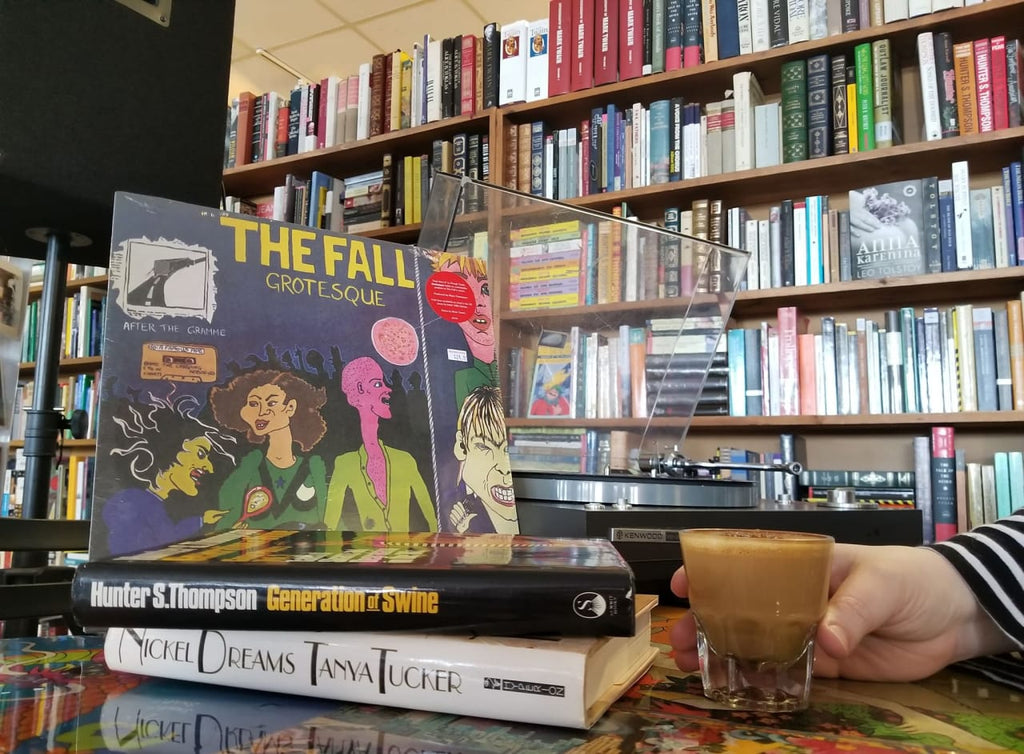 Photo of a record player, a record, a stack of two books, and a hand holding a cortado. 