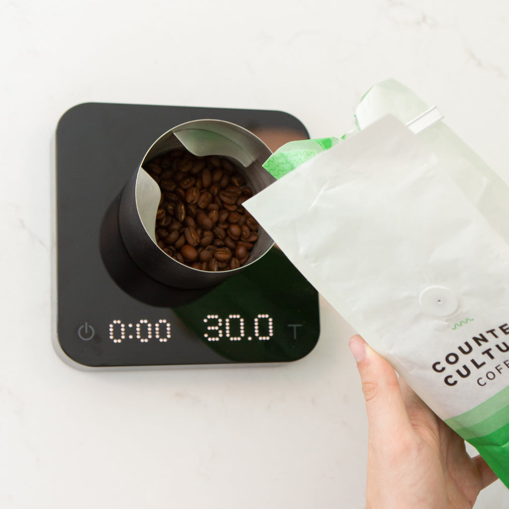 Photo of a person pouring coffee from a bag into a cup for grinding on a digital scale.