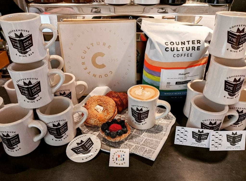 Photo of a cappuccino, baked cooks, a bag of Hologram Counter Culture Coffee, and stacks of mugs that say, "coffeehouse & wine bar. The Den" on them.