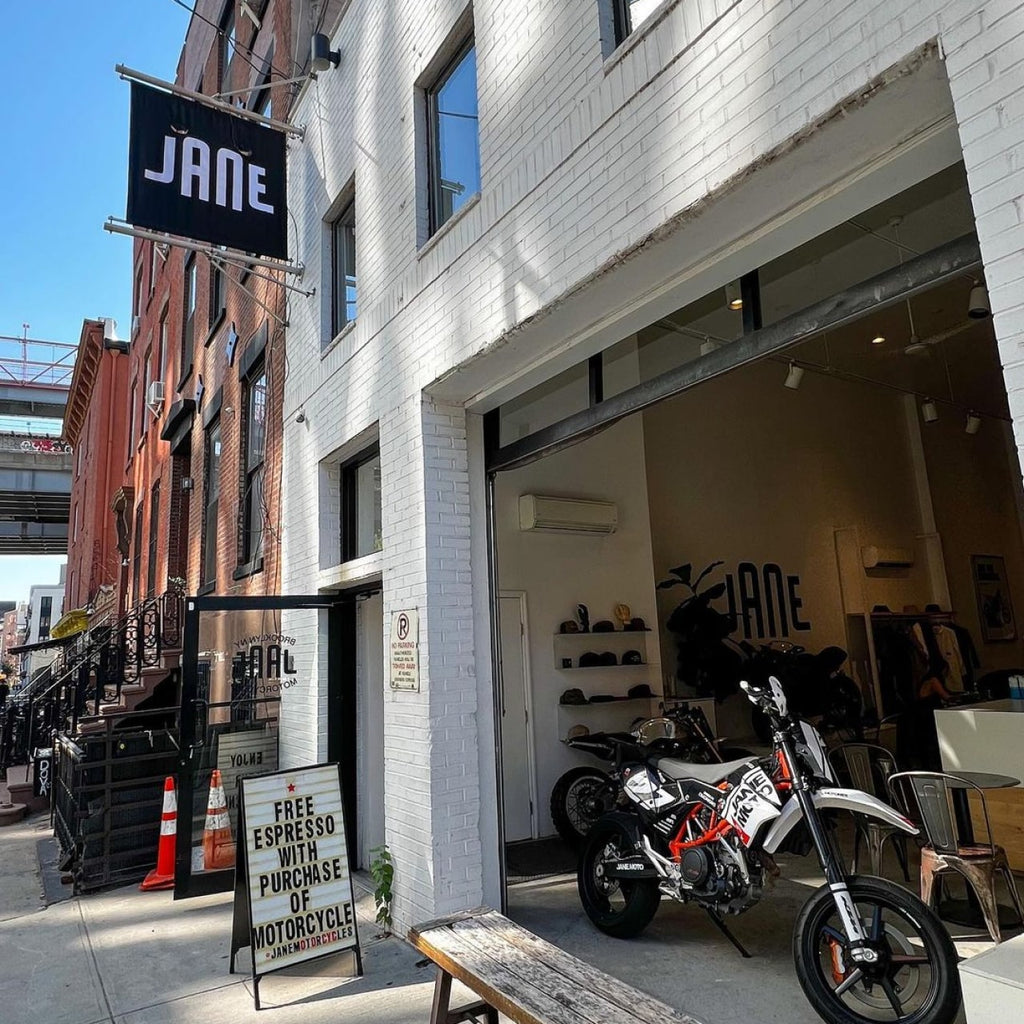 Photo of the exterior of Jane motorcycles. The garage door is up on the brick building and there is a sandwich board sign that says, “Free espresso with purchase of motorcycle.”