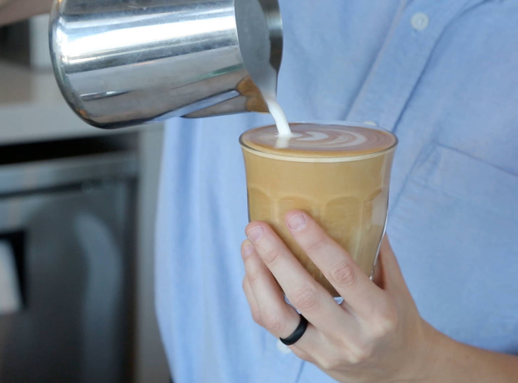 Photo of a person pouring steamed milk from a pitcher into an espresso drink.