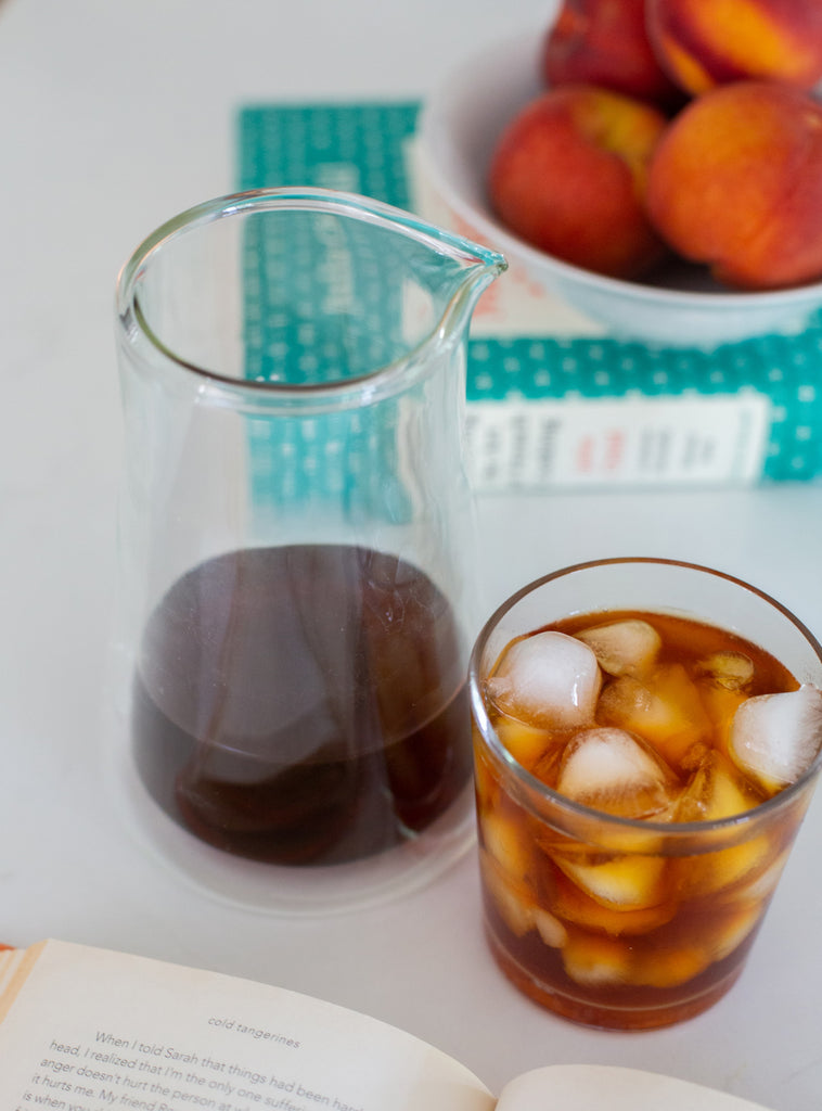 Photo of a carafe of cold brew next to a glass of iced coffee on a counter. Beside them are a book with peaches stacked on top.