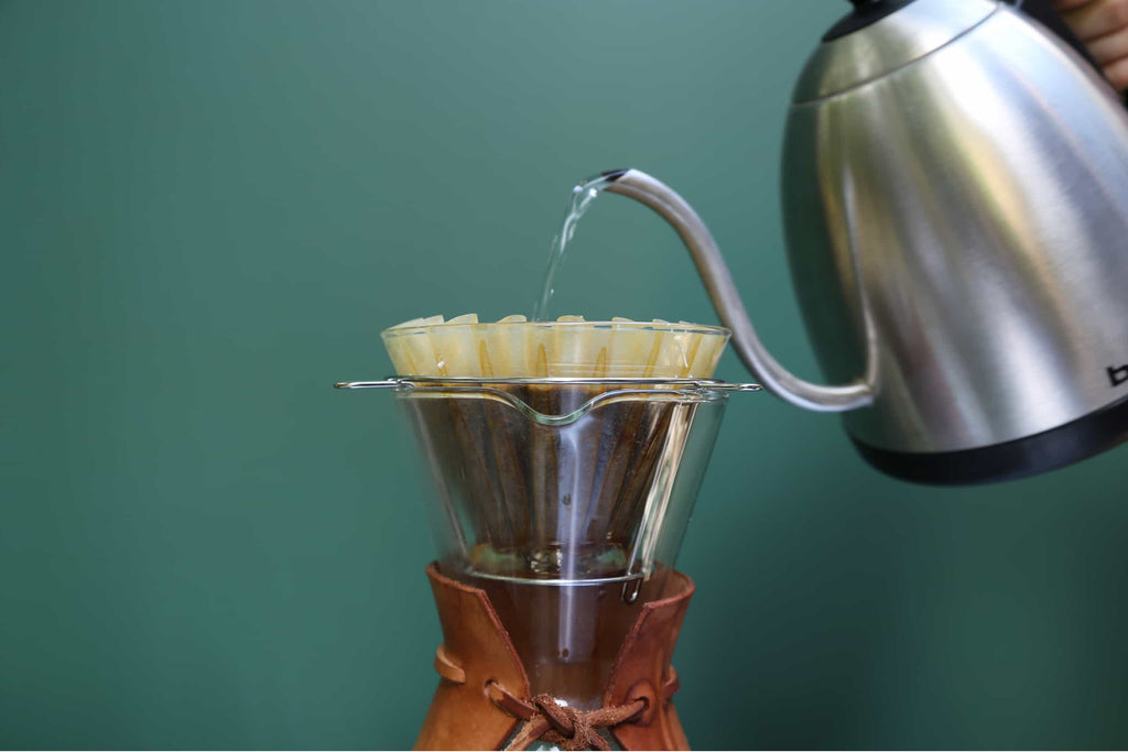 Photo of a person pouring water from an electric kettle over coffee grounds in a pour over device.