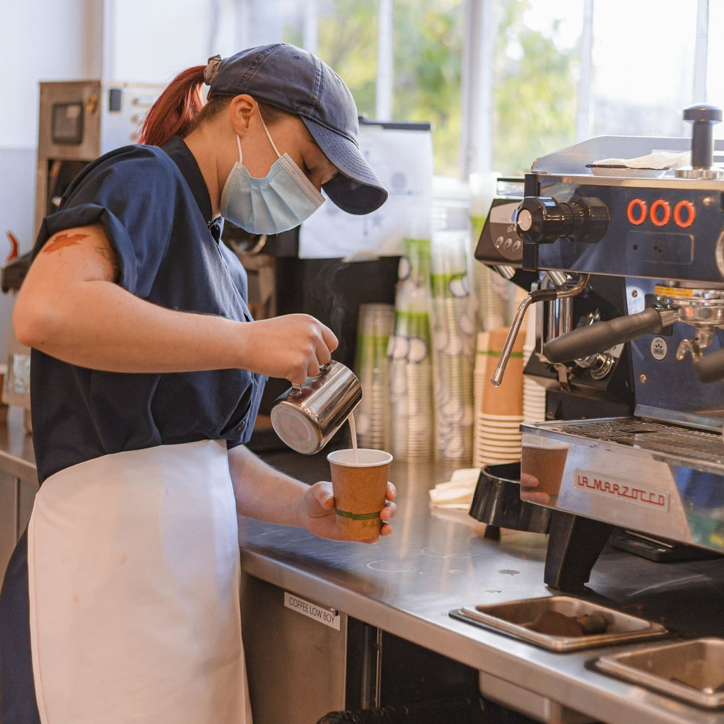 Photo of a person in a mask behind a bar pouring an espresso-based drink.