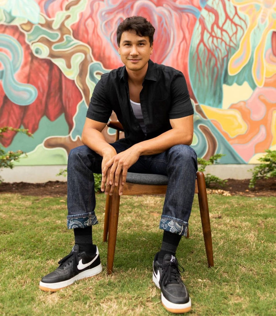 Photo of Gabriel Eng-Goetz sitting on a chair in front of one of his murals and looking at the camera.