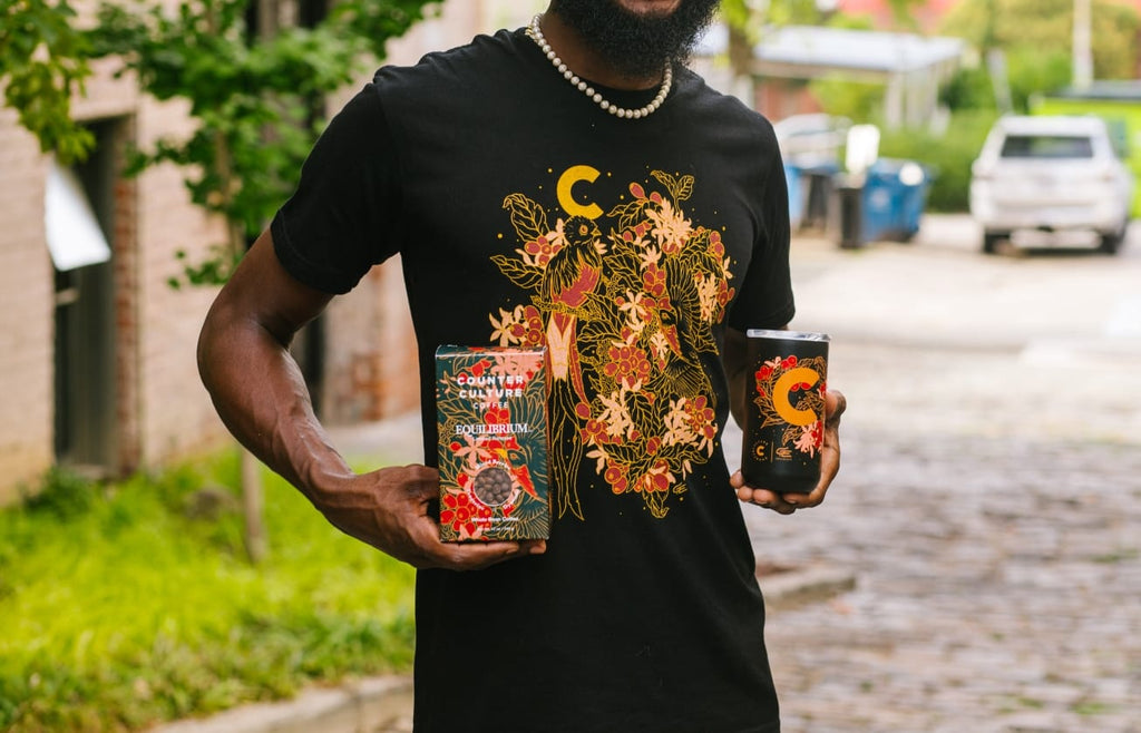 Photo of a person wearing an Equilibrium shirt and holding a box of Equilibrium coffee and the Equilibrium coffee tumbler.