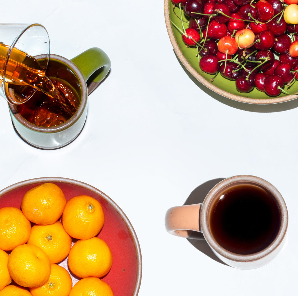 Top down photo of coffee being poured into a mug from a carafe, a bowl of cherries, a bowl of clementines, and another mug of coffee.