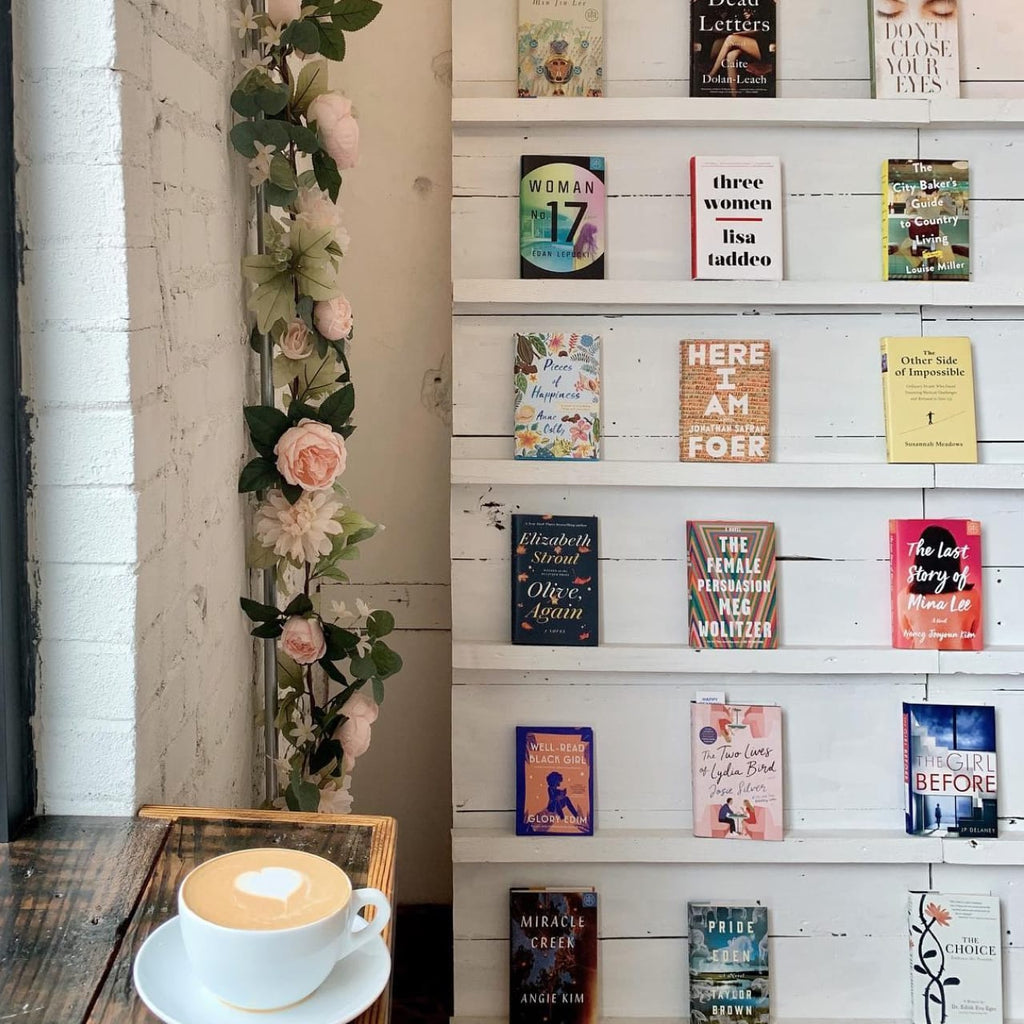 Photo of a window counter with a latte on it. Behind that, there is a bookshelf with an artful display of titles. 