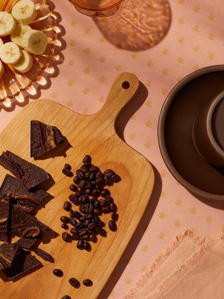 A broken coffee bean and banana topped chocolate bar on a wooden cutting board next to some coffee beans. There is also a pink glass plate with fresh sliced bananas and a brown ceramic plate and coffee mug.