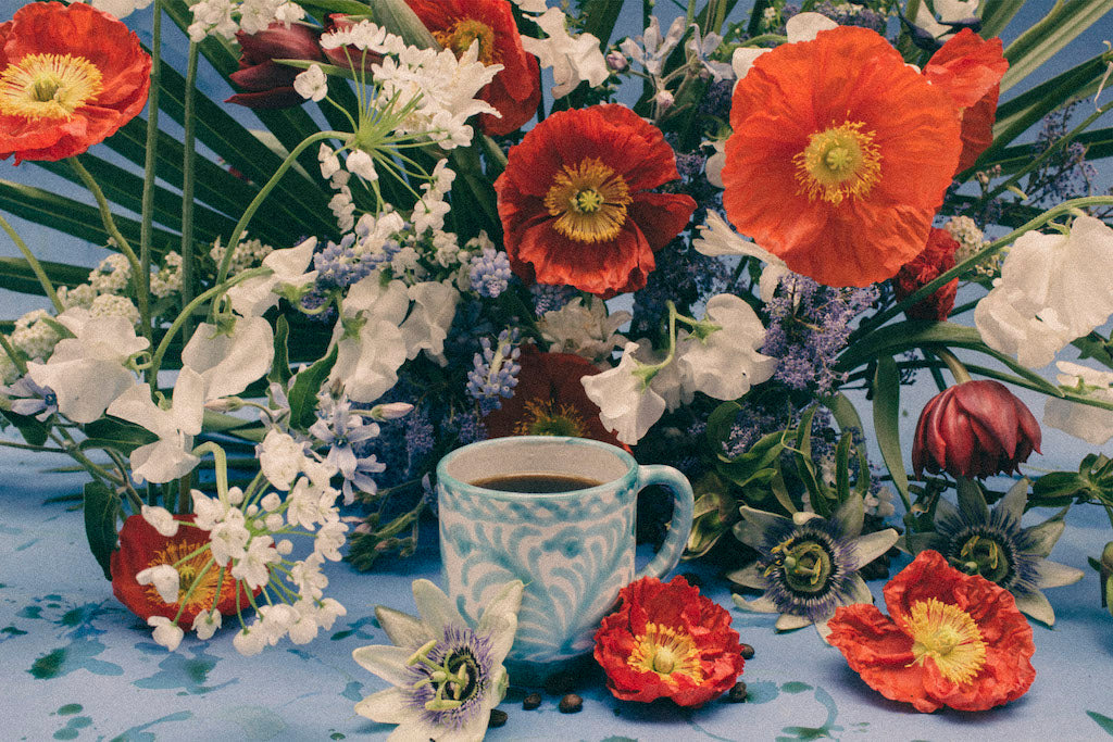 Photo of a mug of coffee in front of a seasonal floral arrangement with sweet peas, poppies, and tulips.