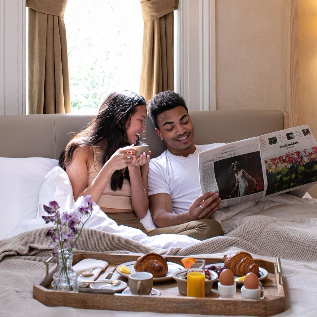 Photo of two people in bed with breakfast and a newspaper.