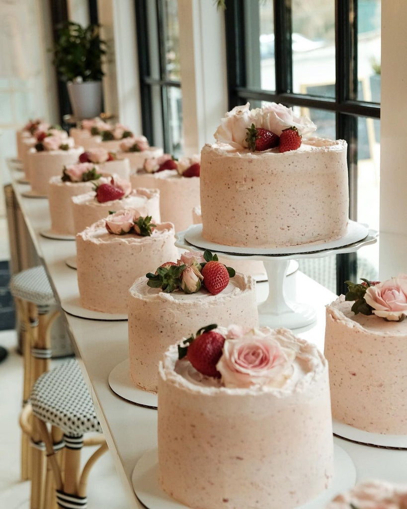Multiple strawberries and cream cakes decorated with berries and flowers lined up on a marble counter.