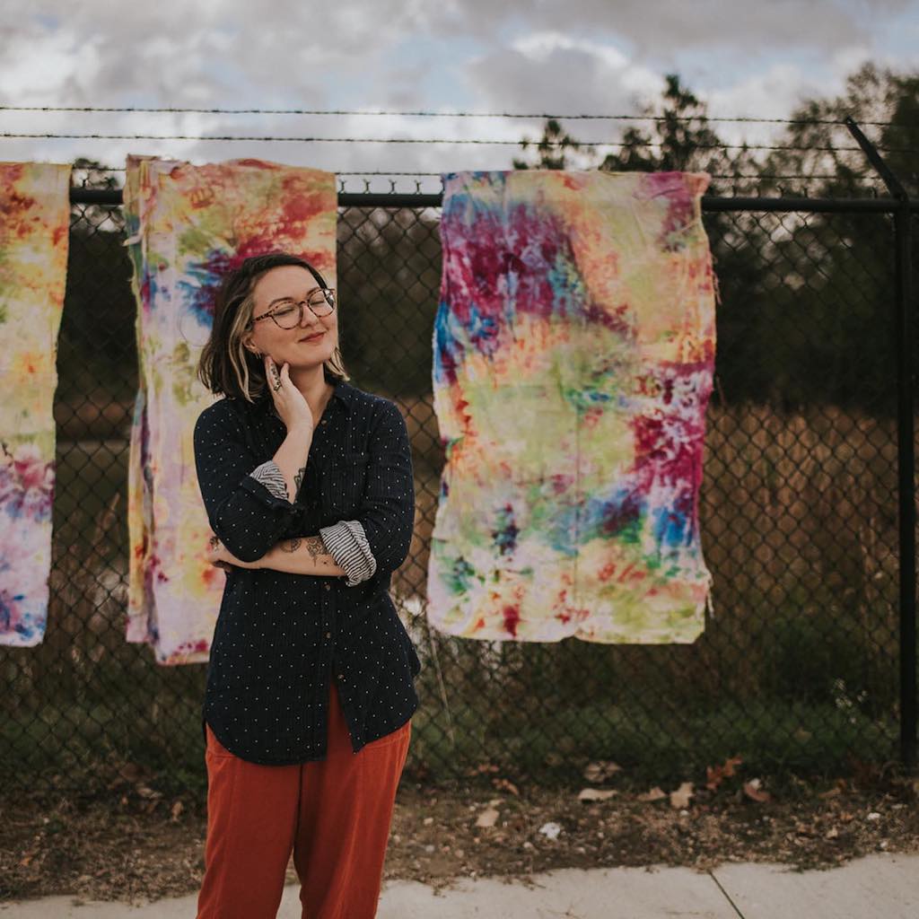 Photo of River standing in front of a fence that has some of their dyed fabrics draped over it.