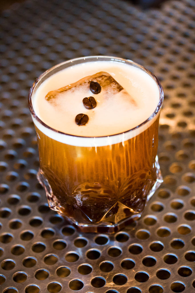 Photo of a rocks glass with a large ice cube inside and a coffee drink. There are three coffee beans lined up on top.