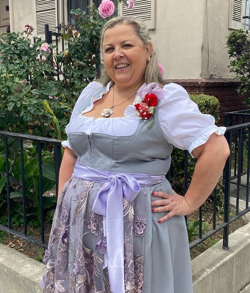 Woman with a plus size figure in a light gray Dirndl with a purple apron, and bow.