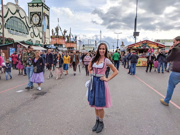 Woman wearing traditional outfit from the alpine regions. A pink dirndl dress with pink dirndl skirt. The skirt length is last her knees. She is wearing a navy blue dirndl apron and cotton white dirndl blouse.