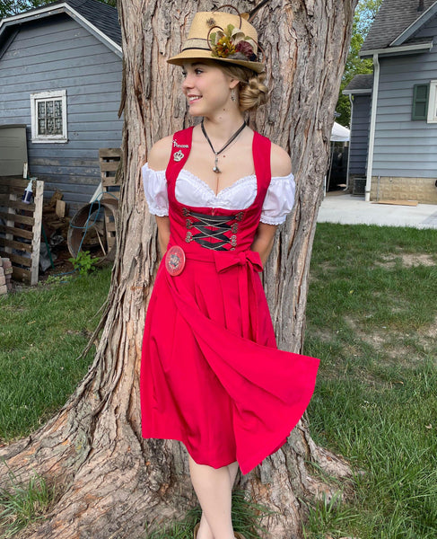 Girl standing in front of a tree, wearing a red dirndl dress, white blouse, and beige hat 
