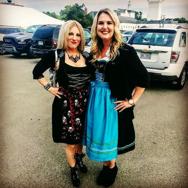 girls wearing black dirndls at a german event. dirndl dress on the left is black with a skull printed apron and the other has a blue apron