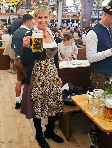 woman wearing a black and leopard print dirndl in one of the beer tents at Oktoberfest. She is holding a liter of beer near a table with food.