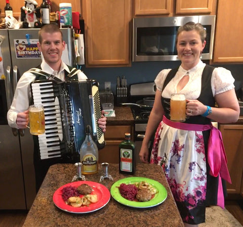 man in his kitchen playing an accordion next to a woman wearing a dirndl with a white dirndl blouse