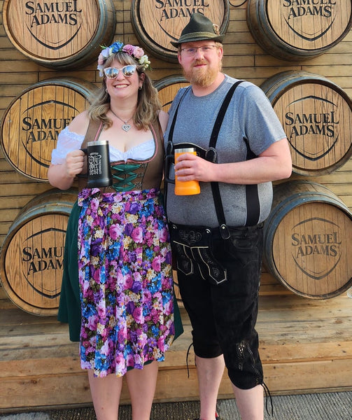 Woman standing in a dirndl, with a floral crown, next to a man with lederhosen, holding beers at an Oktoberfest
