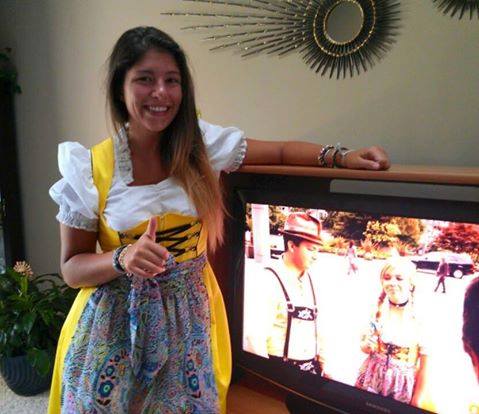 girl wearing a yellow dirndl with a white dirndl blouse standing next to the tv playing the nickelodeon movie swindle with the same dirndl on actress jenette mccurdy