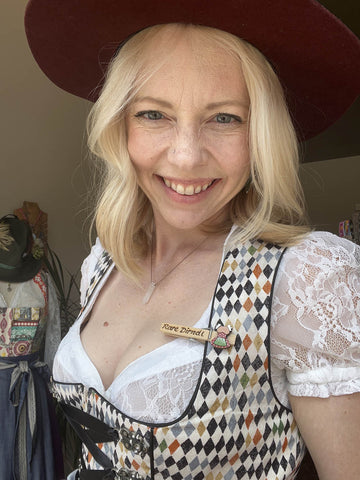 blonde woman wearing a white lace dirndl blouse and a modern dirndl dress smiling and showing her Wiesn Grupperl 