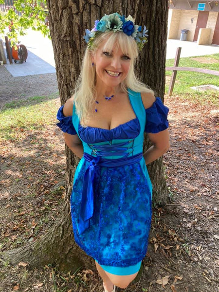 Woman smiling and standing in front of a tree in a blue dirndl with a floral crown 