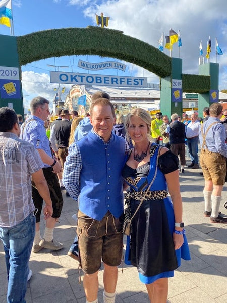 Couple standing under the entrance of Oktoberfest, wearing lederhosen and a german dirndl dress with a tight bodice 