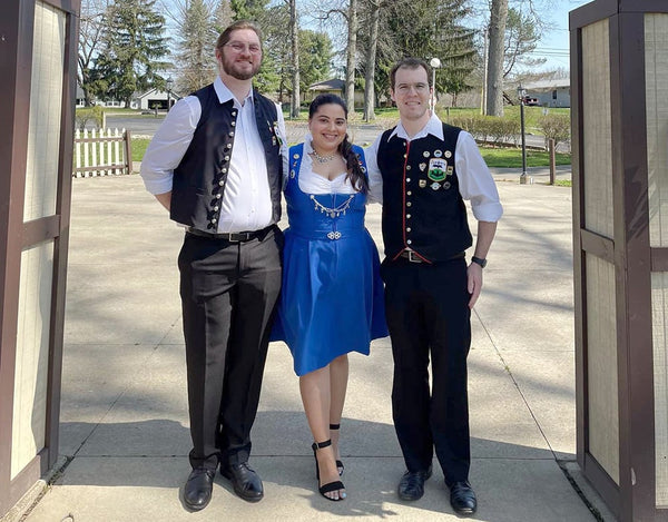 Woman with dark hair in blue dirndl standing between 2 men in black pants and vests. 