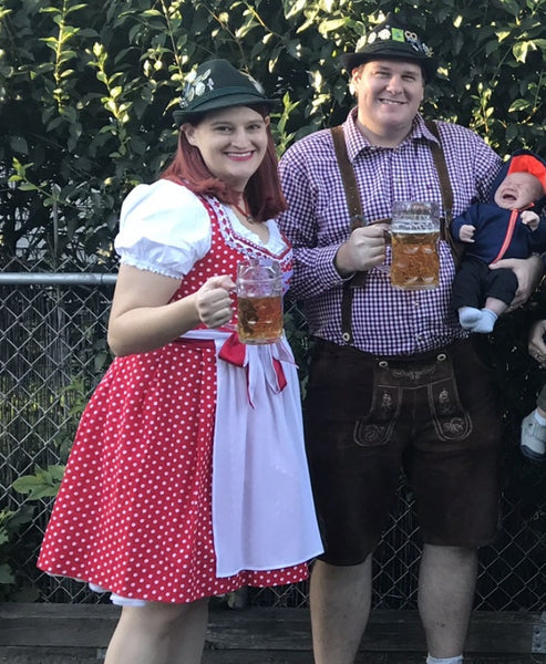 Woman in a red polka dot Dirndl holding a beer with a man in lederhosen and a little baby celebrate in their backyard