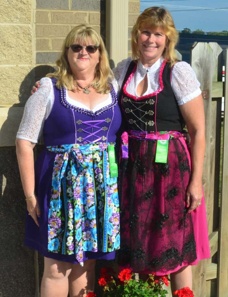 Two women in oktoberfest dirndls with white blouses and aprons. One is in a white cotton blouse with quarter sleeves, purple dirndl bodice with silk lace up and apron with colours. Second woman. is in white blouse with black bodice and hot pick detailing with black lace apron with embroidery.  with short sleeved blouses and colorful aprons 