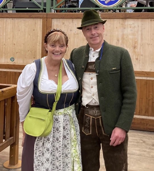 woman wearing a plus size dirndl dress at oktoberfest with a lace apron standing next to a man wearing lederhosen and a trachtenjacke