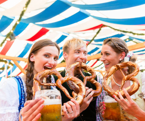 three people wearing dirndl and lederhosen at oktoberfest eating giant pretzels