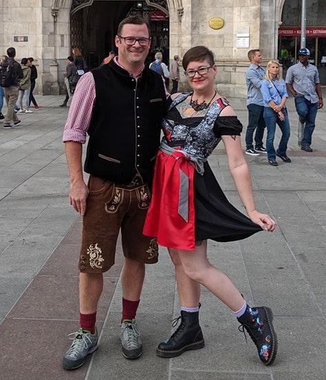 Couple in Munich town square. Women is wearing a black dirndl dress, black blouse, red apron and lace up black shoes. Man is wearing a black vest and lederhose